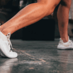 Osoba u iskoraku nosi bele minimalističke patike na podu teretane.